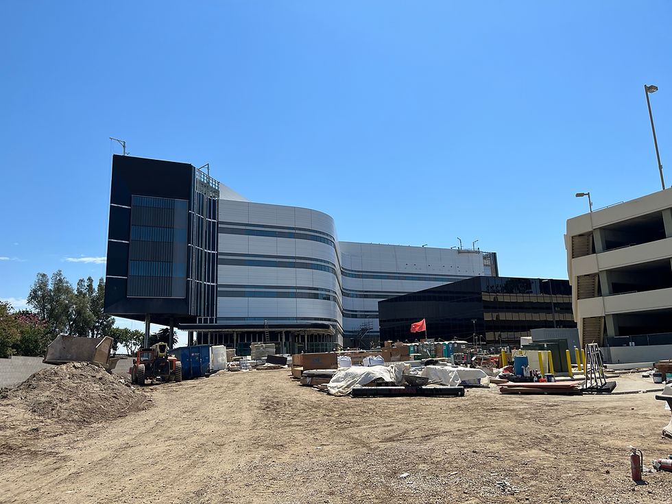 Building glass façade with wave architecture, ground is dirt with building materials, port-a-potties, machines, and yellow safety bollards. Background is blue sky with a couple white thin clouds and green trees. To the right is existing building and parking structure