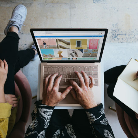 Three people collaborating; one typing on a laptop displaying colorful images, another taking notes in a notebook, and one sitting nearby.