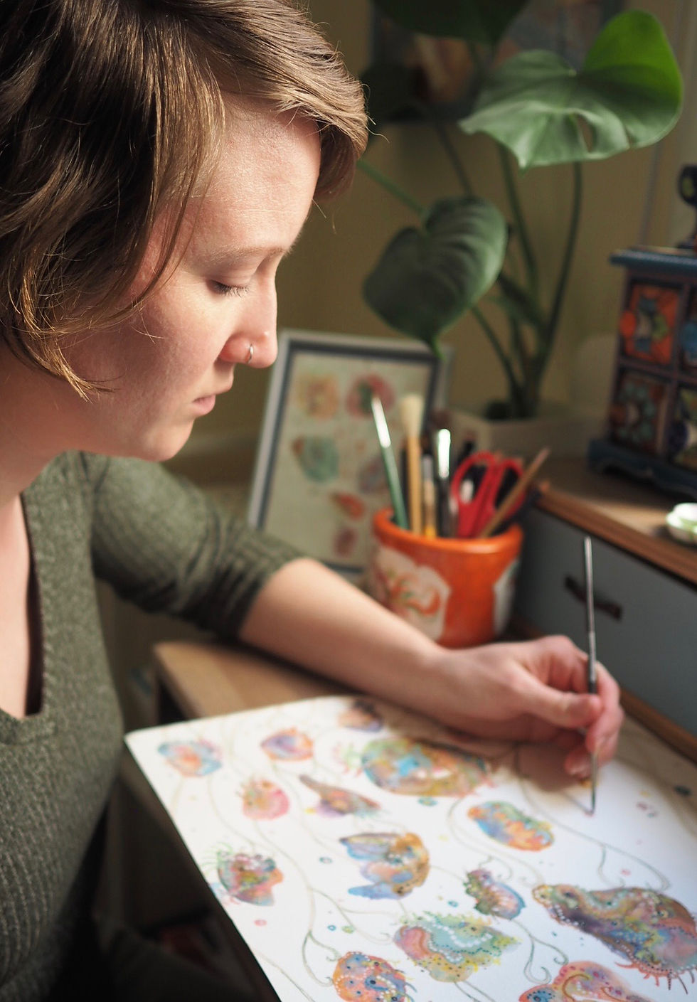 Artist Miriam Rowe from WanderingModa, painting colorful abstract shapes on paper, sitting at a desk with brushes and plants nearby, focused expression.