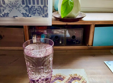 Handmade Roman mosaic coaster beside a purple glass and potted plant on a desk.