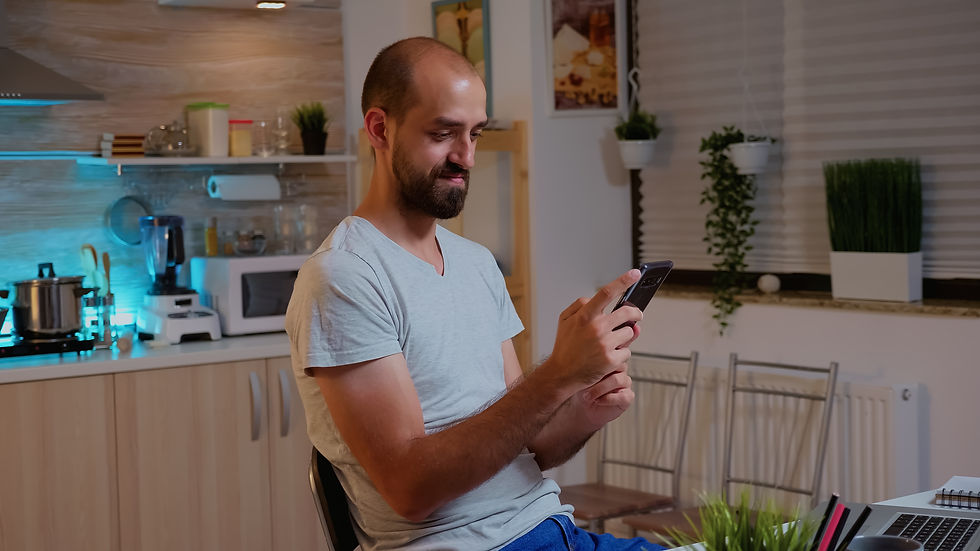 Homem jovem com barba, sorrindo enquanto usa um smartphone em uma cozinha moderna à noite. Ele está sentado em uma mesa de trabalho improvisada com um notebook ao lado.