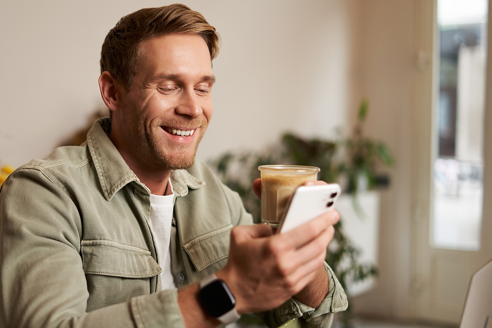 Homem jovem sorridente, vestindo jaqueta verde, segurando uma xícara de café com uma mão e usando o celular com a outra. Ele demonstra satisfação e tranquilidade em um ambiente iluminado e moderno.