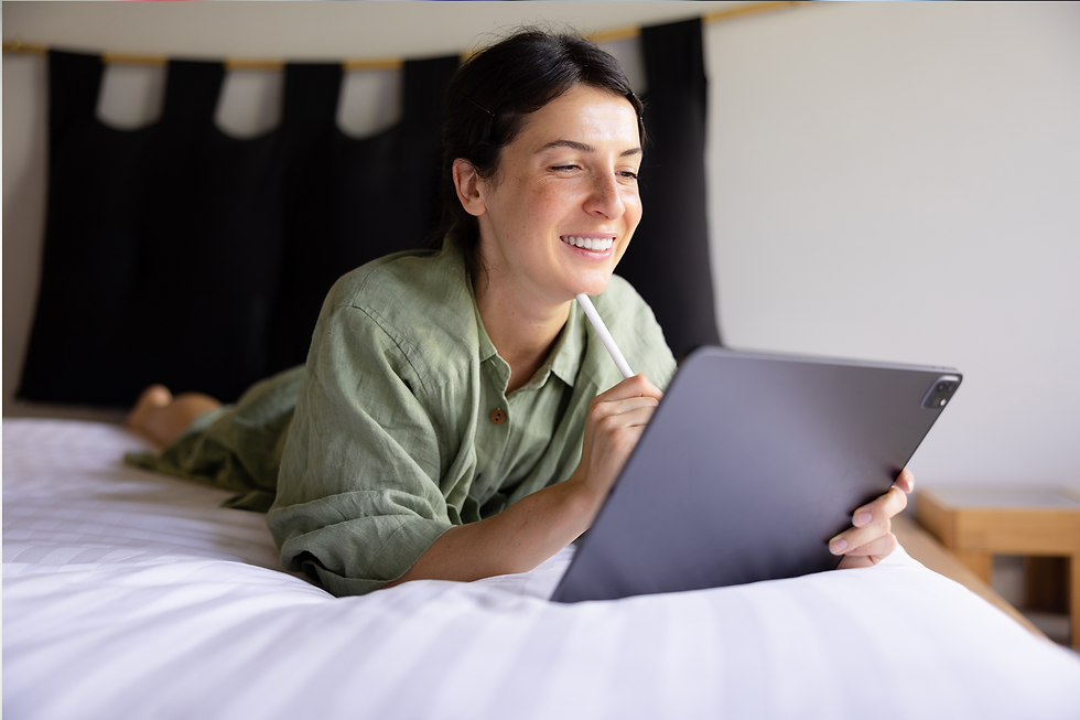 Uma mulher sorridente com cabelos escuros, vestindo uma camisa verde-clara, deita-se de bruços em uma cama com lençóis brancos listrados. Ela segura um tablet cinza-escuro com as duas mãos e usa uma caneta stylus na mão direita, interagindo com a tela. Ela está olhando para o tablet com uma expressão alegre e pensativa. A cabeceira da cama tem detalhes em preto e madeira.