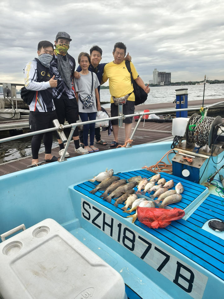 Full outdoor shot of a group of people on a light-blue fishing boat. 

The boat is a small, light-blue, open vessel with a flat deck. A number plate, SZH1877B, is prominently displayed on the deck. Several fish are laid out on the deck, various white and light-colored fish. A red fish (possibly a snapper or similar species) is also visible in the center of the catch.

Five people are standing on the boat’s deck, positioned near the front edge. They appear to be Asian or East Asian, and are dressed in casual clothing. The clothing colors include black, gray, dark yellow-green, and a yellow shirt. Some individuals are wearing jackets or vests on top of their shirts. One person is wearing a gray-colored neck gaiter. They are smiling or have expressions that suggest a happy occasion.

Behind the boat, the background shows a body of water, with a hazy cloudy sky visible; it appears to be a calm day at sea. A series of industrial grain silos are barely visible in the background on the water, along the very edge of the horizon line. 

The overall tone of the image is one of a relaxed, outdoor activity, likely a fishing trip. The lighting suggests a daytime shot, and the weather is clear but not sunny. The focus is on the boat and its occupants, with the background being somewhat out-of-focus.