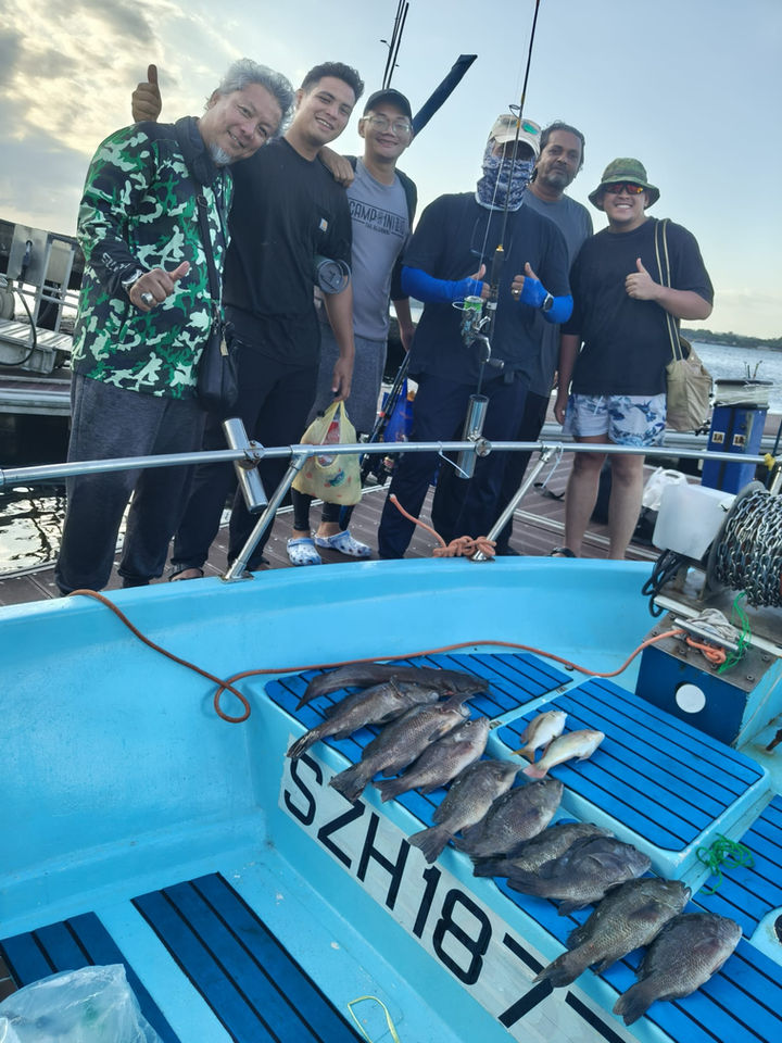 A group of men stand on the deck of a light-blue fishing boat, smiling and posing for a picture. 

The men are diverse in appearance. Some are wearing camouflage-patterned shirts, while others have on dark-colored, casual tops. One man is wearing a light-colored baseball cap. Several of the men are holding fishing rods and equipment. 

On the boat's deck, a considerable number of freshly caught fish are arranged and visible. The fish are dark grayish-black in color, and they rest on the deck, with various sizes and positions. 

The boat's deck is a light-blue, with horizontal dark-gray stripes running along it. A registration number or code, "SZH1877", is clearly visible on the boat's deck, slightly below the level of the men.

The background includes a partly cloudy sky, suggesting an outdoor setting. A portion of the shore and some trees or vegetation can be seen in the distant background.  The overall impression is of a successful fishing trip.