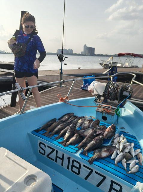 Full shot of a person on a small, light-blue fishing boat. 

A woman with shoulder-length brown hair and sunglasses is standing on the boat's deck, holding a small, dark-colored rectangular bag. 

Numerous fish are laid out on the boat's deck, which features a blue-striped, non-slip surface. The fish appear to be a mixture of dark and light varieties, likely caught during a fishing trip. The larger fish are clustered in the front, middle portion of the boat. Smaller, lighter-colored fish are scattered behind and around the larger fish filling the space.

The boat's deck is light-blue, and a license plate, marked with the characters "SZH 1877", is visible.

A fishing rod and reel are partially visible on the boat's deck, along with a chain-like apparatus possibly used for hauling in larger catches. On the background is a body of water with some buildings and a small vessel on the far right of the frame. The scene suggests a sunny day at a marina or waterfront area. The background shows a cityscape in the distance.