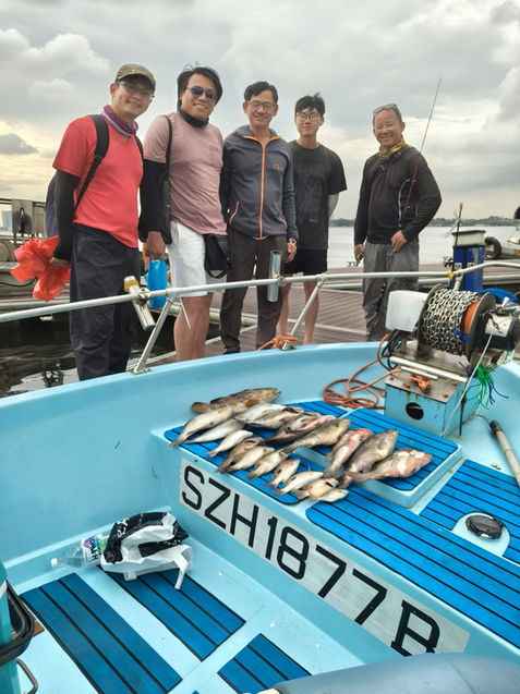 A group of men stand on a light-blue boat deck, likely after a fishing trip. 

Four men are positioned on the deck. The man on the far left is wearing a reddish-pink long-sleeved shirt and khaki-colored pants. The man next to him is wearing a light pinkish-tan top and white-ish shorts. The man in the center is wearing a dark-grey top and pants with a dark blue/gray t-shirt underneath. The man on the far right is wearing a dark navy/black colored top and pants. All are wearing baseball caps or similar head covering.

Several caught fish are laid out on the boat's deck. The deck is light blue with dark blue/black stripes making up a pattern. The letters "SZH1877B" are visible, possibly indicating the boat's registration or identification number, displayed prominently on the deck. Various accessories including a fishing reel are also visible on the boat deck. 

The background suggests an outdoor setting, likely a lake or a similar body of water, with cloudy skies. The water surface is partially visible in the image.