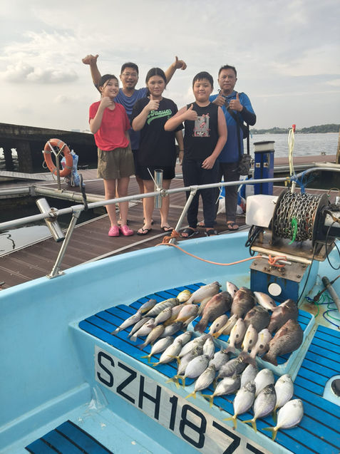 Full shot of a group of people on a light blue, motorboat-style vessel. 

The foreground shows the deck of the boat, a light teal-blue color, with several levels of wooden slats creating a raised, flat surface.  A large quantity of various fish are laid out on the flat surface, in different sizes and shades of gray, silver, and light brown -- characteristic of caught fish. A license plate, SZH1877, is clearly visible on the middle front part of the deck. 

In the mid-ground, a group of approximately five people are standing on the front part of the deck. They are diverse in ethnicity and age, and all are smiling broadly, demonstrating joyful engagement.  Some of them are holding up their thumbs, suggesting a celebratory aspect. Two men and three youths, seemingly between 10 and 17 years old, are positioned somewhat tightly, facing somewhat towards the camera. Their clothing is a mix of casual shirts, t-shirts, and shorts.

The background hints at an open water scene, with a partly cloudy sky. Some light gray/blue water is vaguely visible, along with the part of a dark dock or pier in the background towards the far side of the scene (the right). The focus is clearly on the boat and the people.