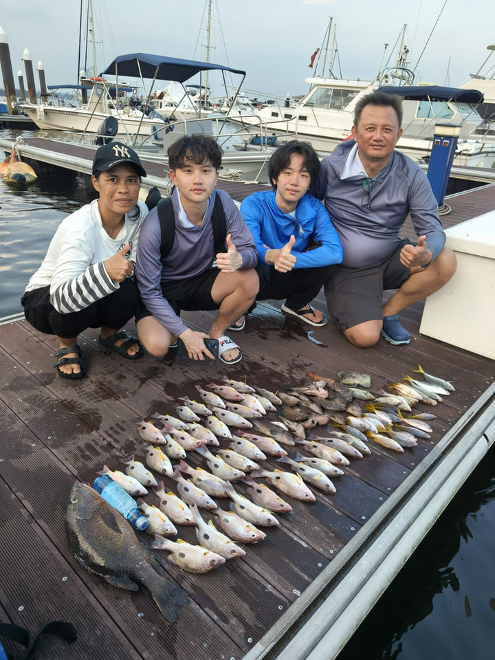 Full outdoor shot of a group of people sitting on a wooden dock, likely after a fishing trip. 

The foreground shows a wooden dock, dark brown and gray in color, with a substantial collection of various fish laid out. The fish are mostly small to medium-sized, light yellow-tan, and light gray/silver in color, showing various species.  A dark-blue plastic water bottle is visible near the middle and lower portion of the image.

Four people are seated, facing the camera. A woman, wearing a white shirt with striped sleeves, is on the left, next to a light-skinned young man in a gray, long-sleeved shirt.  Immediately to his right is a similar-looking light-skinned young man in a blue long-sleeved shirt, and slightly further to his right, on the right side of the image, is a light-skinned older man in a gray, long-sleeved fishing shirt, seated.  All are crouched down. The people are smiling and appear to be enjoying themselves.

The background shows a marina or dock area with various boats and motor vessels of varying sizes and styles docked. The boats are primarily light cream, tan, or white in color. The sky is a light grayish-blue, indicating a day without intense sunlight. The lighting suggests an overcast or partially cloudy day.