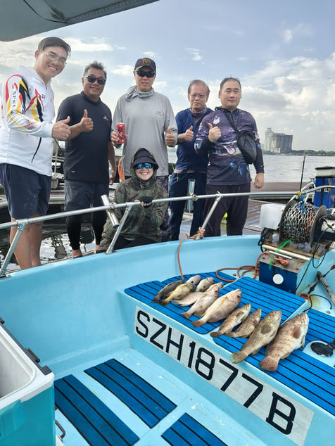Full indoor shot of a group of people on a light-blue fishing boat, likely in a harbor or bay. 

Several men and one woman are standing. The men are positioned closely together, wearing casual dress attire, including fishing tops and shorts. Some are wearing baseball caps, and all are smiling and giving thumbs-up. A soda can sits between the two men in the middle.

The boat's interior is light-blue, with blue-and-white horizontal stripes on the interior seat and steps that run along the bottom, a registration plate or number, "SZH 1877 B", is visible on the boat's center console. Several fish are prominently displayed on the bottom of the boat. The fish are various species and sizes, lying on the surface of what looks like a hard, laminated mat.

The background is out-of-focus, suggesting a coastal scene.  A partly cloudy sky and a distant water horizon can be seen. A portion of a building or structure is visible in the distance, indicating a cityscape or potentially an industrial zone.

The overall impression is a joyous group celebrating a successful fishing trip aboard a small fishing boat.
