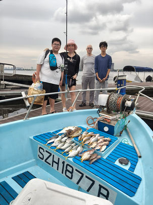 Full outdoor shot of a group of people on a boat, likely after a fishing trip. 

Five people stand on the deck of a light blue/gray-colored small fishing boat. The passengers are positioned in front of the boat's cockpit. Three of the individuals are young men, one is a young woman, and one is a man of middle age. The woman is wearing a light pink/rose-colored hat. They are mostly dressed in casual, light-colored clothing. Visible items suggest they are involved in fishing; fishing rods and a tackle bag are visible. A quantity of freshly caught fish are prominently displayed on the boat's seating areas, indicating a successful fishing trip. The fish appear to be various species of small to medium-sized fish, piled on top of each other. The boat's interior is light blue with dark blue/black horizontal seating/storage compartments. The boat's hull is a lighter shade of bluish gray. 

The background shows a partially visible pier or dock with a section of water, and the sky is mostly overcast with a light gray tone. A variety of items, including other boats, are partially visible in the distance. The overall impression is of a group of people enjoying a day of fishing together.
