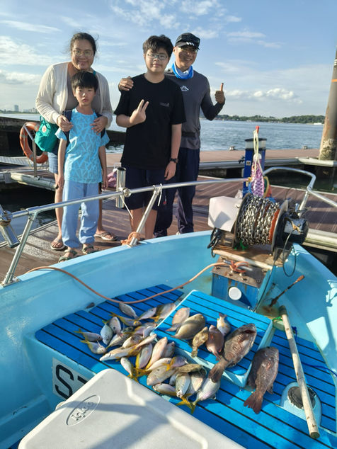 Full indoor shot of a family of four on a boat, likely fishing. 

The foreground shows a light-blue boat with a blue-striped, wooden-looking deck. On the deck, many smaller fish (likely different types of fish) are piled and spread out. A few larger, darker fish are scattered among the smaller ones. The deck looks like it is part of the boat's interior. 

In the mid-ground, a family of four is standing on the deck. A woman and a teenage boy are in the center of the group.  A middle-aged man is standing next to the boy, while another teenage boy is beside the man. The adult woman and the teenage boy are wearing clothing of light-colored colors, likely made of cotton, and the adults appear to be casual or semi-formal attire. The boy on the right appears to have darker clothing resembling a t-shirt or a loose top, and the man on the right is wearing a darker shirt along with a neckerchief. They all have a natural stance and appear to be engaging in a happy or relaxed activity. 

The background shows a body of water, with a calm blue/gray surface. The sky above has a light and partly cloudy appearance. Sections of a pier, or possibly a dock, are visible along the water's edge, just beyond the end of the boat. Lighting is natural and bright, implying an outdoor setting. A fishing reel and equipment are seen on the top part of the boat, and fishing poles are present.