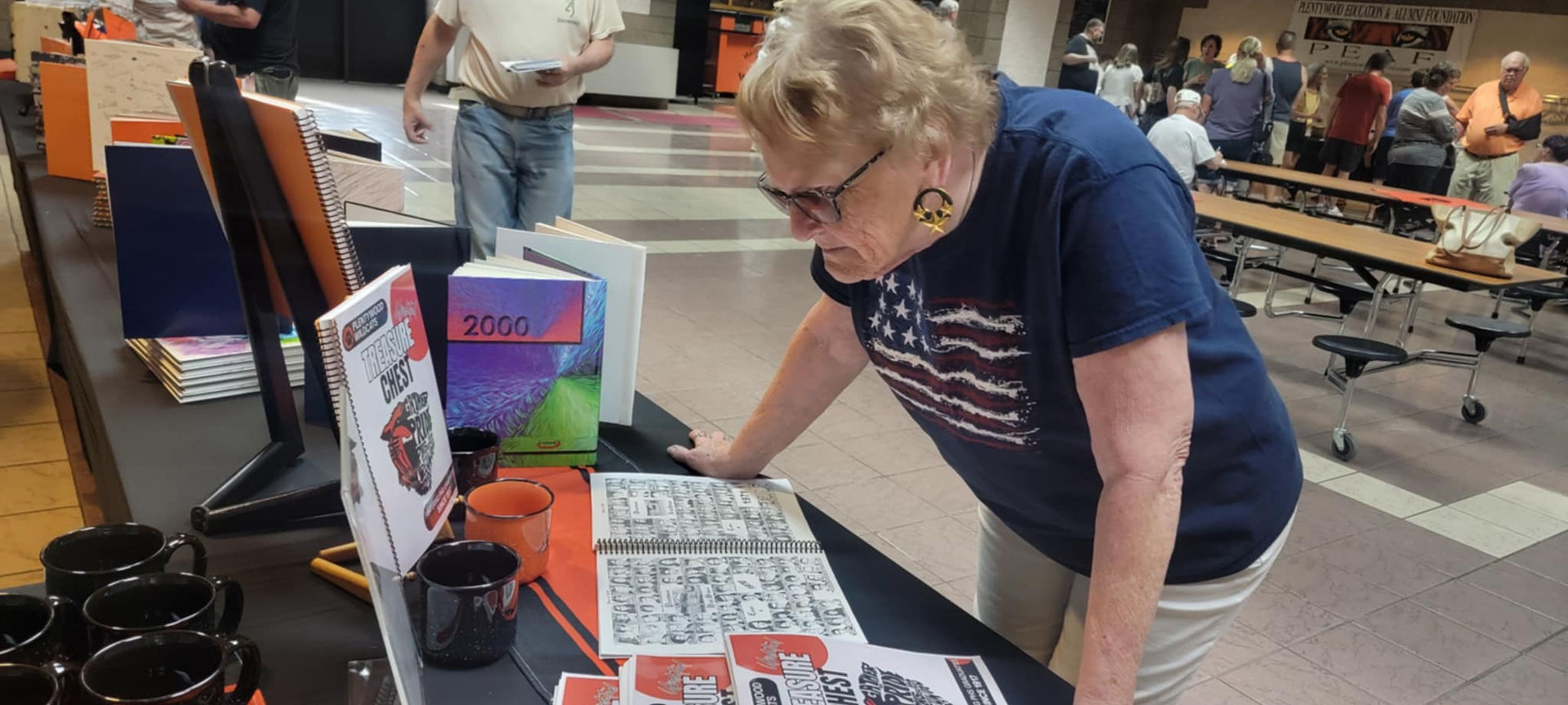 Woman examines items at a table displaying printed materials and mugs during an event.