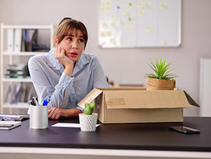 A woman thinking on the desk. She has box with stuff from her desk. She has been made redundant.
