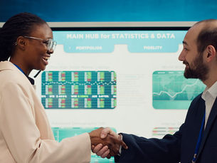 A business manager and new employee share a warm, confident handshake during a data briefing meeting. A Black woman wearing glasses and a beige blazer smiles at her colleague, a bearded man in a dark navy suit. A large screen displaying financial statistics, portfolio charts and market data fills the background. Paula Donnan Advisory.