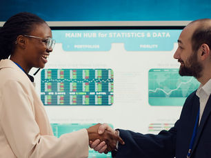 A business manager and new employee share a warm, confident handshake during a data briefing meeting. A Black woman wearing glasses and a beige blazer smiles at her colleague, a bearded man in a dark navy suit. A large screen displaying financial statistics, portfolio charts and market data fills the background. Paula Donnan Advisory.