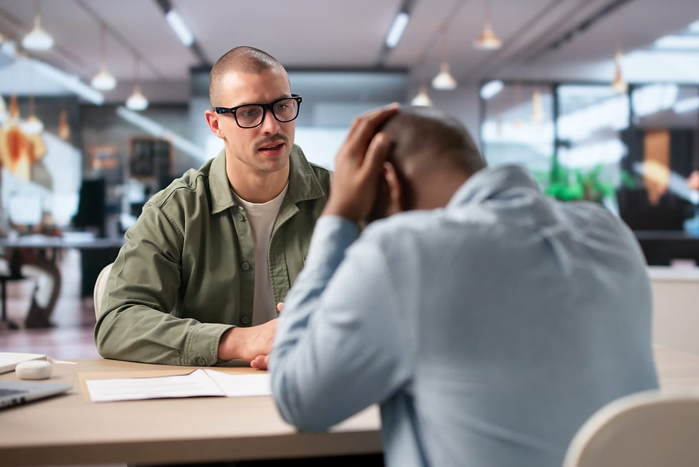 Two professionals sitting across a table in a modern office. One man faces the camera with a concerned expression. The other has his head in his hand, suggesting frustration or difficulty during a workplace conversation. Paula Donnan Advisory