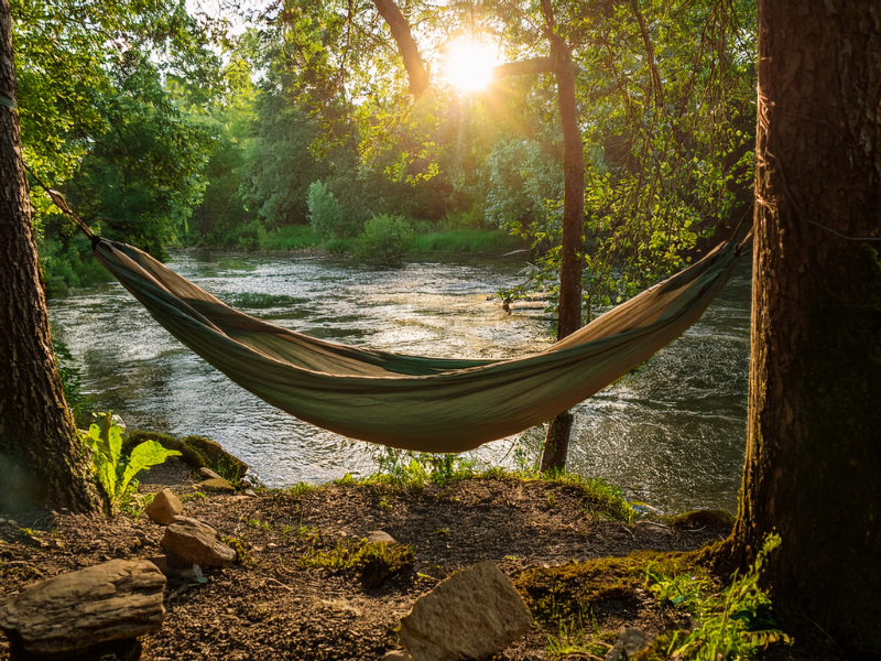 A hammock hangs between trees by a river at sunset. Sunlight filters through leaves, creating a peaceful, warm atmosphere.
