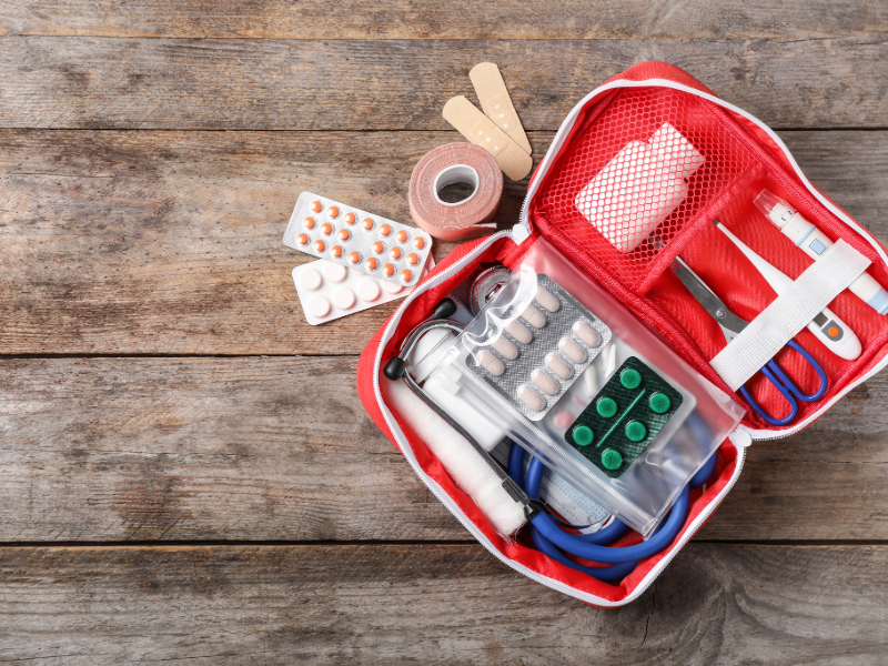Open red first aid kit on wooden table, containing scissors, pills, bandages, and medical supplies. Neutral background, organized setup.