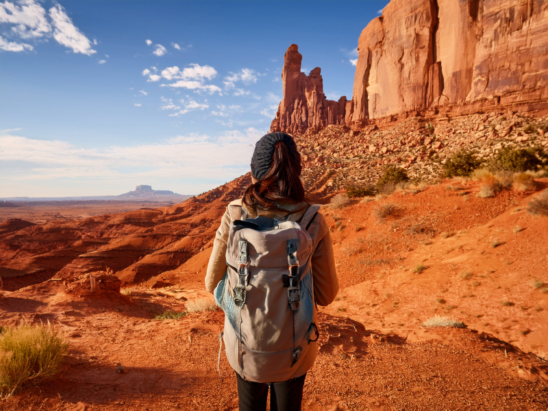 A person with a backpack stands on red rocky terrain, gazing at towering formations under a blue sky with clouds, suggesting adventure.