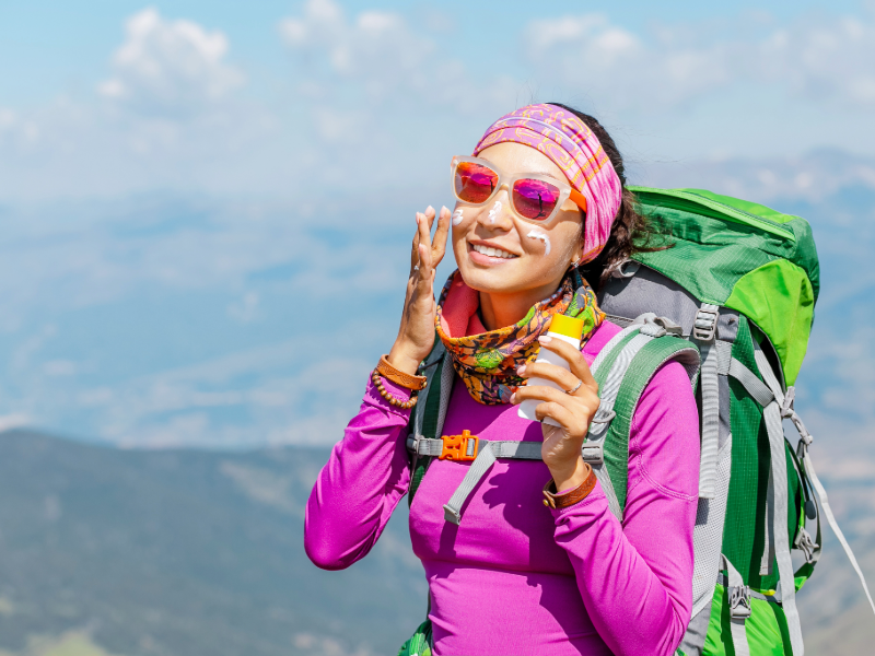 Woman in pink top hiking, applies sunscreen, smiles with joy. Wears sunglasses, headband, and green backpack. Mountainous background, clear sky.
