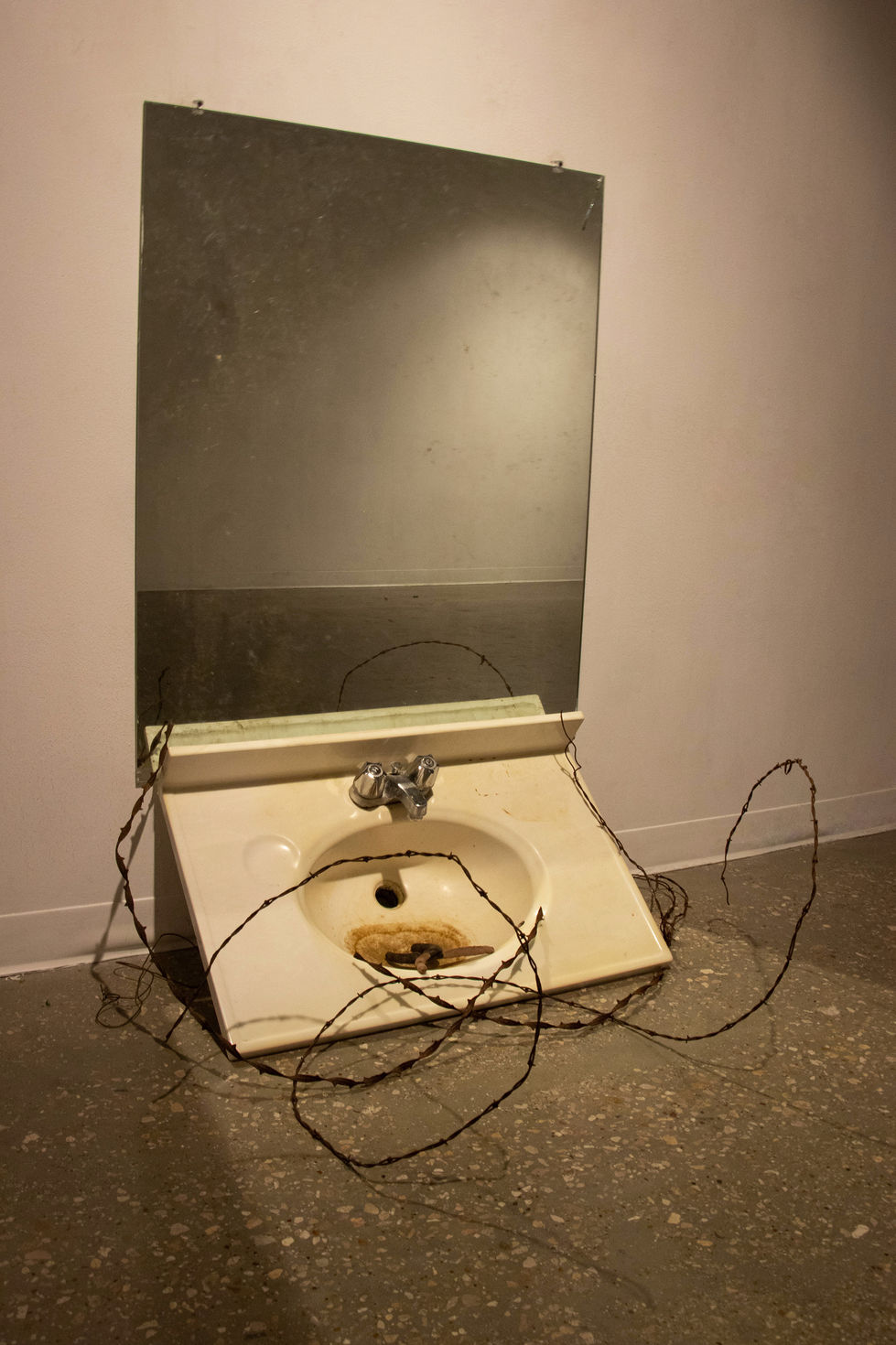 Sink is resting against the wall. Mirror is hanging above the sink. Sink is dirty and covered in barbed wire and pliers.