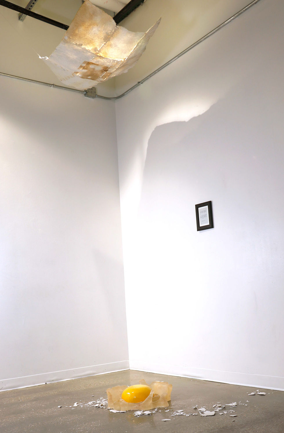 Hanging paper drawing hovering over broken egg sculpture. There is a small framed poem on the wall.