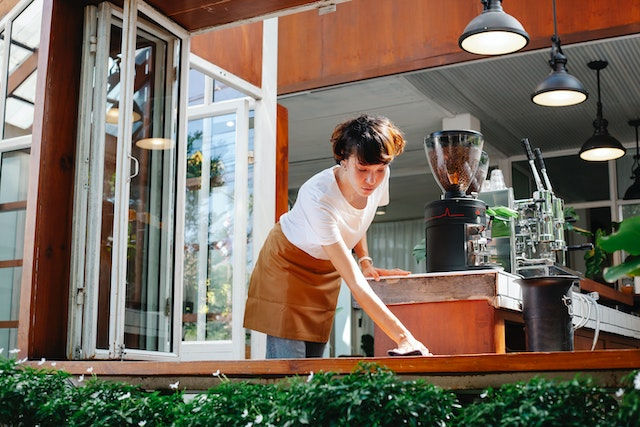 A female barista is cleaning the window sill of a cafe. The window is the size of the wall and has plants underneath it. The barista has short brown hair and is wearing a white shirt and brown apron.