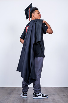 Senior boy wearing a graduation cap, holding a gown over his shoulder, standing on a wooden floor