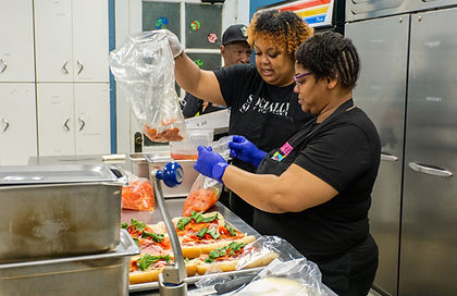 Two people in black aprons work in a kitchen; one person teaches the other how to assemble an Italian sandwich.