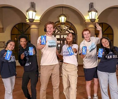 Group of six smiling students holding blue letters spelling “THANKS” while posing together in a warmly lit arched hallway.