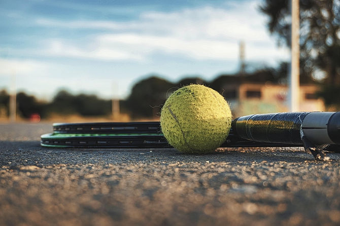 Close-up of a tennis ball resting on the ground beside two tennis rackets, with a blurred outdoor background.