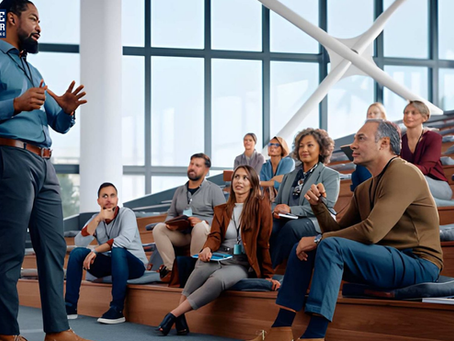 Man in blue shirt speaks to engaged audience seated on tiered steps in modern conference room. Large windows in background.