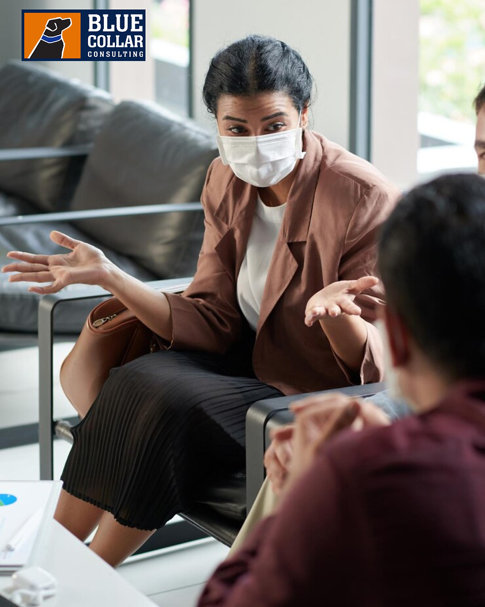 Woman wearing mask gestures while talking with others indoors. She sits on a couch with logo "Blue Collar Consulting" in corner.