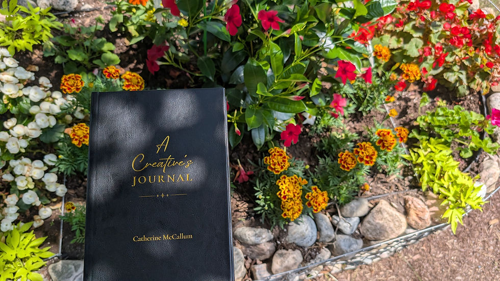 A Creative's Journal, a black hardcover journal with gold text, sits in front of a flower garden with a range of red, orange and yellow blooms