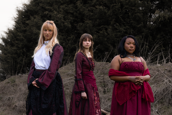 Group portrait of three women in red and burgundy gowns standing together in an open field.