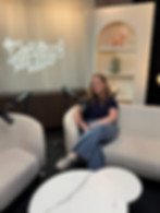 Olivia Mae Hanlon sitting on a white bouclé sofa in a modern podcast studio. Behind her, a neon sign glows with the words 'The Girls in Marketing Podcast.' The studio features professional microphones, a marble coffee table, and an arched shelving unit with decorative art.