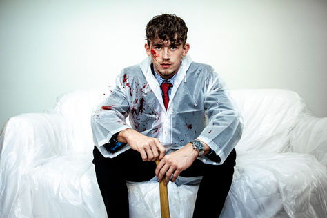 Male model seated on a white sofa wearing a transparent raincoat over a shirt and tie, holding an bloody axe, American Psycho inspired fashion shoot