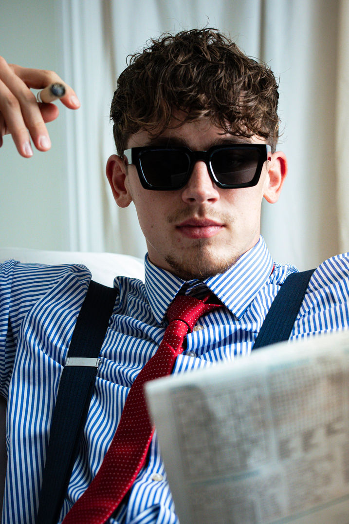 Male model wearing sunglasses, shirt and suspenders, reading a newspaper, American Psycho inspired menswear shoot