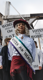 Documentary-style photograph of suffragette reenactors holding “Votes for Women” protest signs.