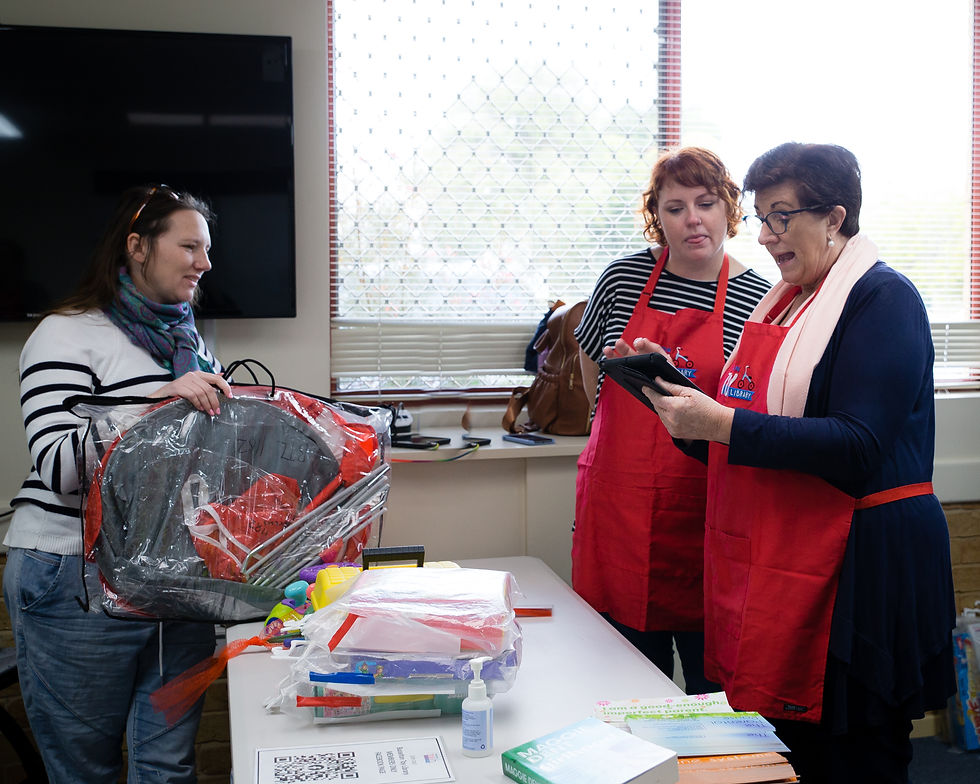 Two adults in red aprons stand behind the toy library loans desk in a bright room, one holding a tablet and another adult stands on the other side of the desk holding up a dress up in a bag ready to be loaned.