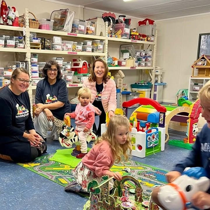 Smiling adults and toddlers play in a bright toy-filled room, with colorful play mats, shelves, and many toys.