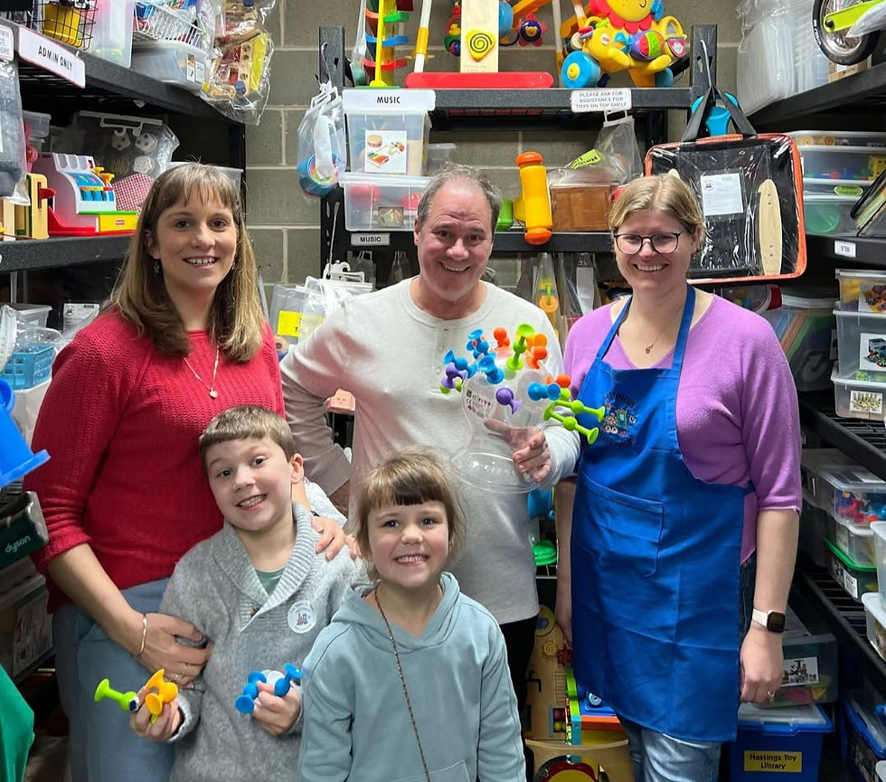 Smiling adults and two children pose in a toy library, holding colorful toys; shelves filled with toys behind them.
