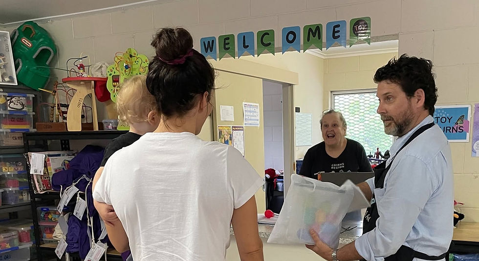 A volunteer carrying a bag of returned toys speaks with a parent holding a toddler inside a toy library while another volunteer smiles from behind the counter. Shelves of toys surround the borrowing area, illustrating a typical toy library session.