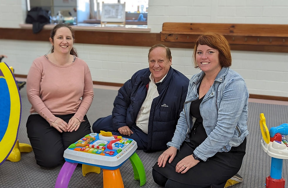 Three smiling adults kneel on a carpet around a colorful toddler activity table in a bright indoor room with white brick walls.