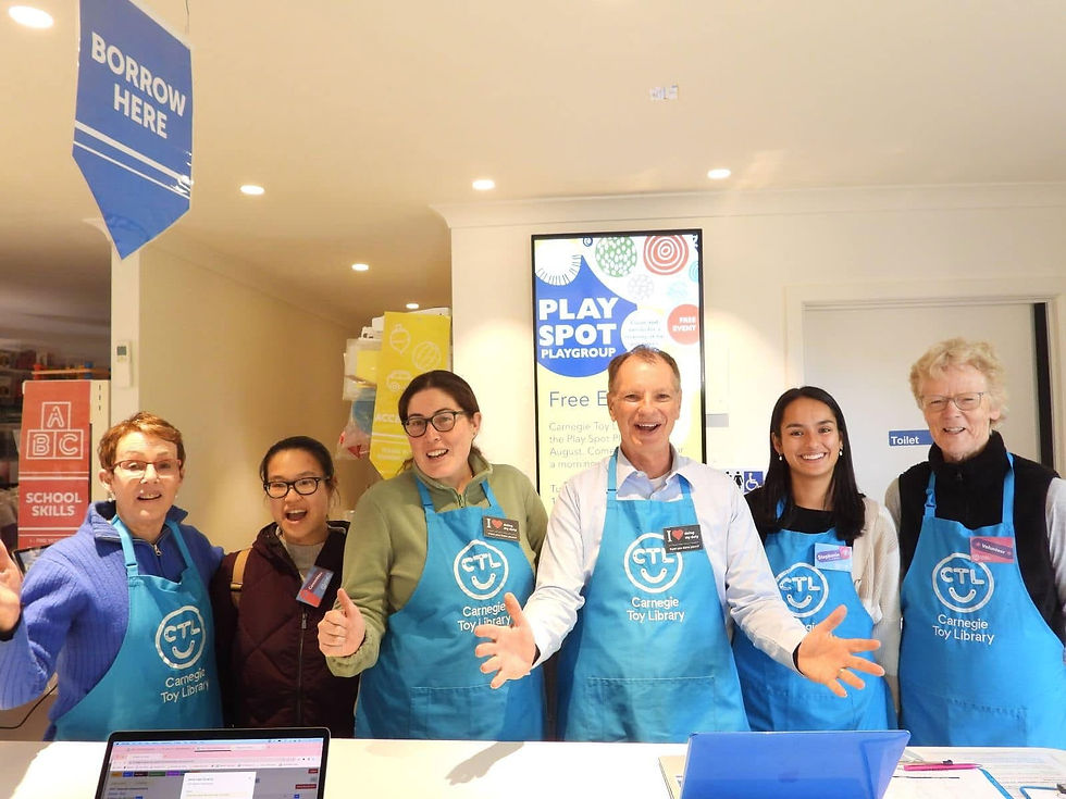 Smiling volunteers and a local MP in blue Carnegie Toy Library aprons stand behind a desk.