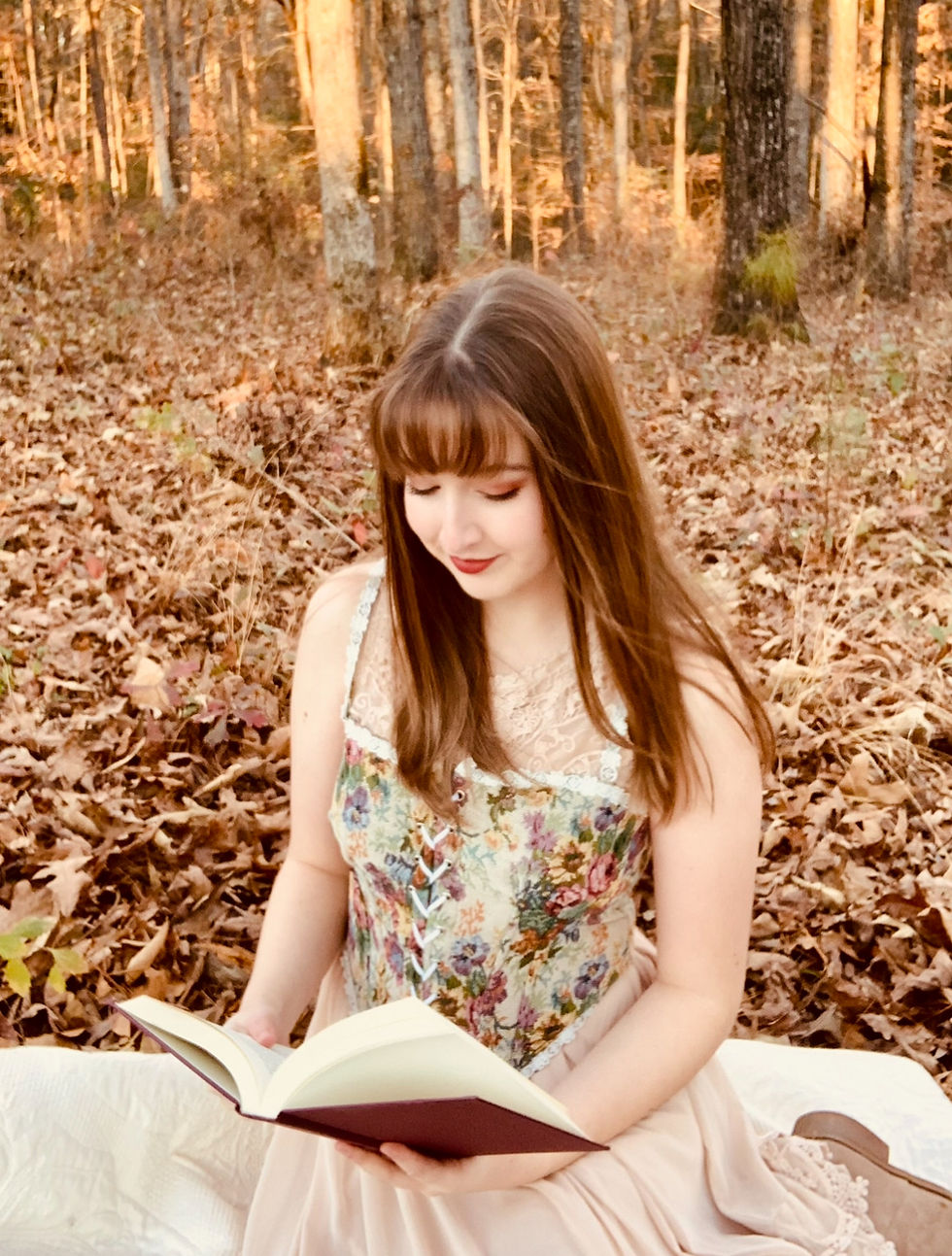 A young brunette woman wearing fantasy-inspired clothing sits reading a book in autumn leaves.