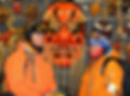 2 skiers in ski gear posing on either side of a large, red, decorative Japanese mask