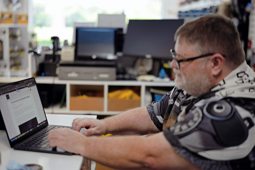 Man with glasses types on a laptop in an office setting, two monitors in background. Wearing patterned shirt, focused and working.