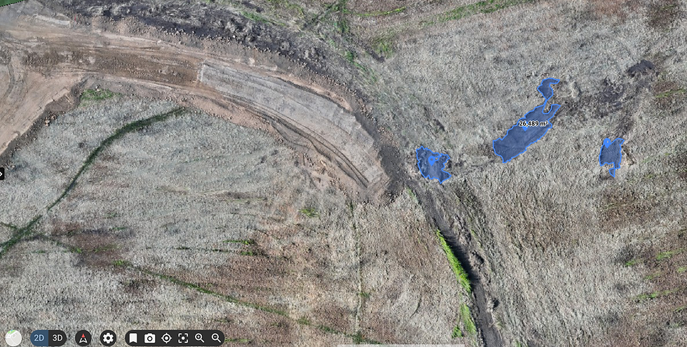 Hochauflösendes Drohnen-Orthofoto (Draufsicht) einer Baustelle mit ersten Erdarbeiten und Fahrspuren. Auf dem unebenen Gelände sind drei spezifische Flächen mit blauen Umrissen digital markiert, wobei eine Teilfläche mit einem exakten Messwert von 26,489 Quadratmetern beschriftet ist.